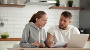 Focused young couple checking analyzing utilities bills sitting together at kitchen table, serious husband and wife reading bank loan documents with laptop, family managing finances planning expenses